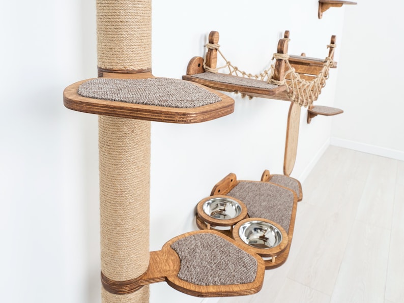 May include: A multi-level cat tree with a sisal-wrapped post, wooden platforms, a hammock, and food bowls. The platforms have a carpeted surface. The cat tree is made of wood and rope, and is designed to be mounted on a wall.