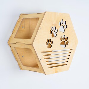 May include: A light-colored wooden cat house with a unique hexagonal shape. The front features paw print cutouts and horizontal slats. The house is designed to be mounted on a wall, offering a cozy space for a cat. The wood has a natural finish.