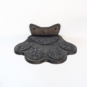 May include: A dark brown, paw-shaped cat shelf with a textured, gray carpeted surface. The shelf has a cat-ear design at the top and is designed to be mounted on a wall. The shelf is made of wood.