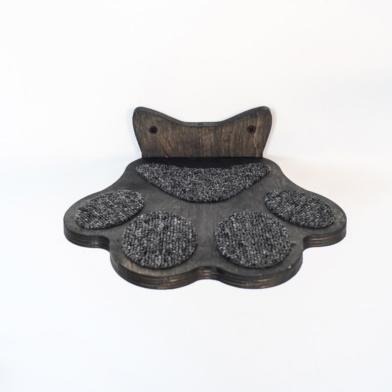 May include: A dark brown, paw-shaped cat shelf with a textured, gray carpeted surface. The shelf has a cat-ear design at the top and is designed to be mounted on a wall. The shelf is made of wood.