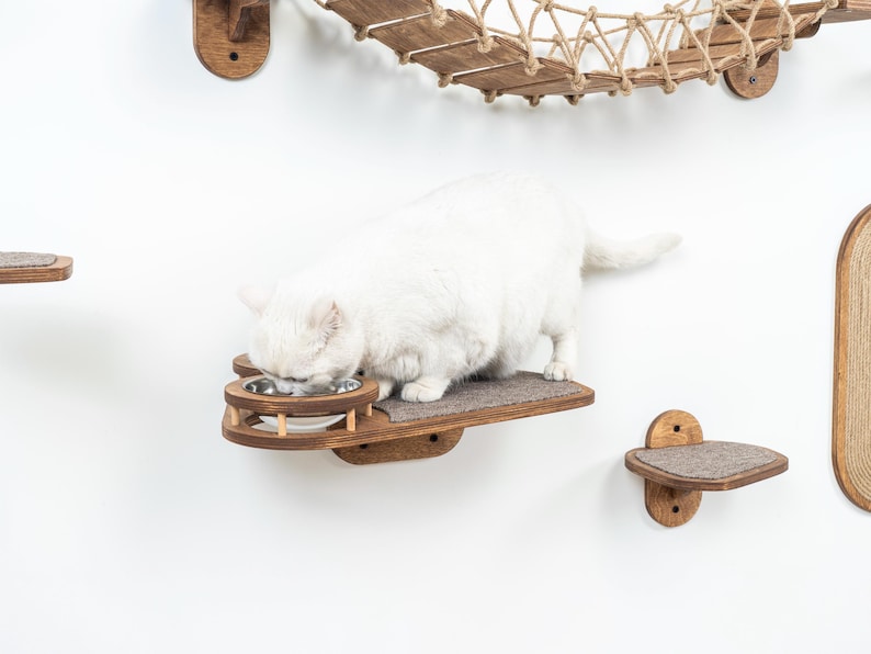 May include: A white cat eating from a stainless steel bowl set into a wooden cat shelf. The shelf is part of a wall-mounted cat furniture system with a rope bridge and other platforms. The cat is on a gray felt pad.