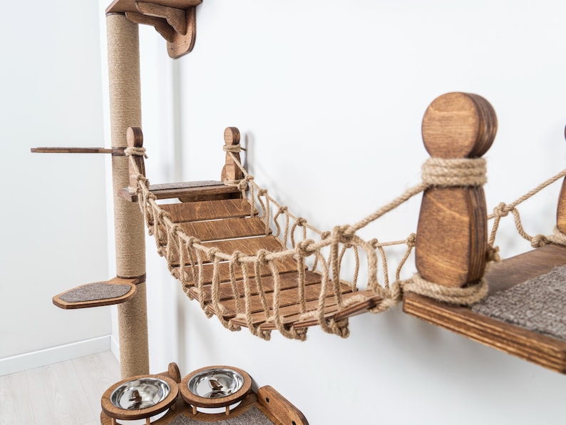 May include: A wooden cat tree with a bridge, scratching post, and platforms. The bridge is made of wooden slats and rope. The scratching post is wrapped in sisal rope. The platforms have a carpeted surface. The cat tree is stained a medium brown color.