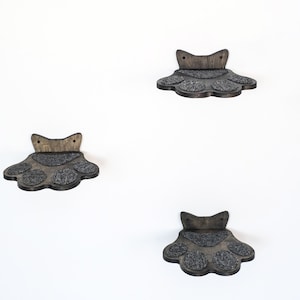 May include: Three wooden cat paw-shaped shelves mounted on a white wall. Each shelf has a dark gray, textured surface for the cat to stand on. The shelves are made of dark brown wood.