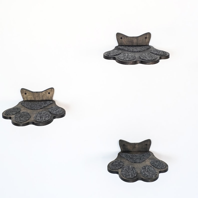 May include: Three wooden cat paw-shaped shelves mounted on a white wall. Each shelf has a dark gray, textured surface for the cat to stand on. The shelves are made of dark brown wood.