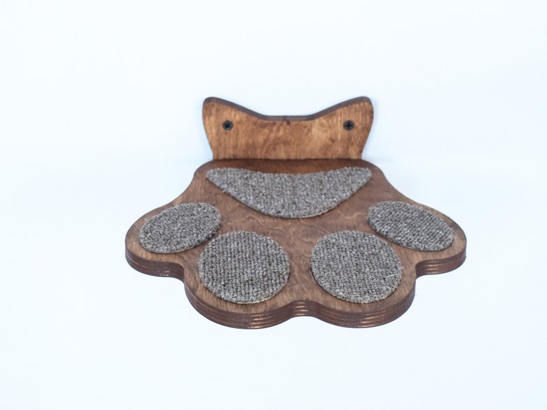 May include: A wooden cat paw-shaped shelf with a dark brown finish. The shelf has four circular and one oval-shaped gray carpeted pads for the cat to rest on. Two screws secure the top portion to the wall.