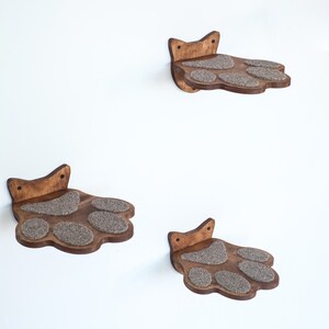 May include: Three wooden cat shelves shaped like paws, mounted on a white wall. Each shelf has a dark brown finish and features gray, textured pads for the cat to rest on. The shelves are designed to be mounted on the wall.