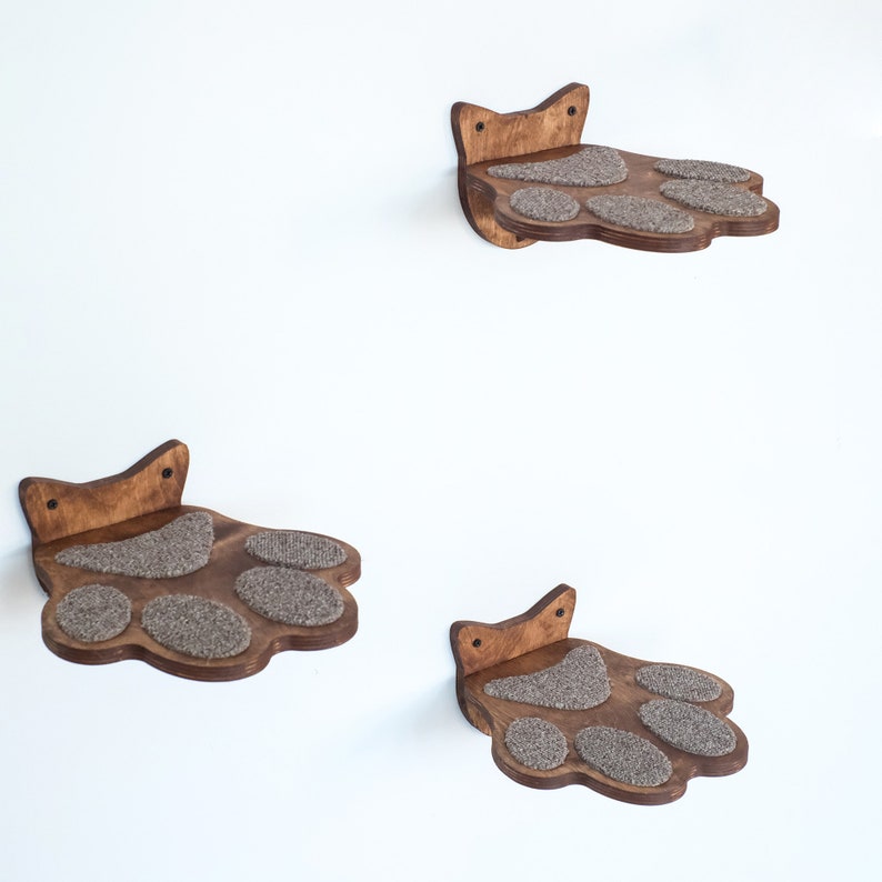 May include: Three wooden cat shelves shaped like paws, mounted on a white wall. Each shelf has a dark brown finish and features gray, textured pads for the cat to rest on. The shelves are designed to be mounted on the wall.