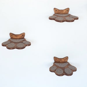 May include: Four wooden cat shelves in a paw print design are mounted on a white wall. Each shelf has a dark brown wooden frame with a light brown, textured surface for the cat to rest on. The shelves are spaced apart.