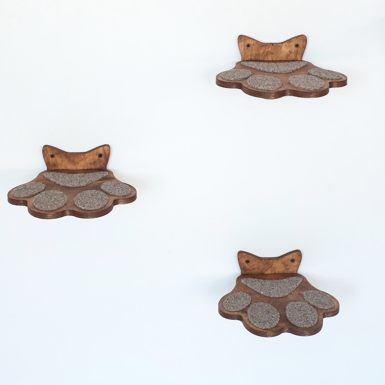 May include: Four wooden cat shelves in a paw print design are mounted on a white wall. Each shelf has a dark brown wooden frame with a light brown, textured surface for the cat to rest on. The shelves are spaced apart.