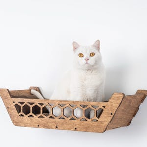 May include: A white cat with yellow eyes sits in a wooden cat bed. The bed is mounted on a white wall and has a geometric honeycomb design. The cat bed is made of light brown wood.