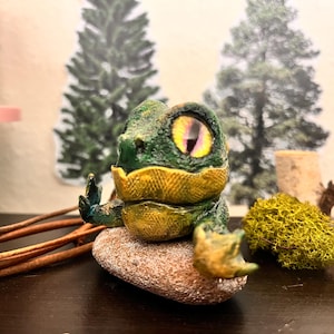 Puede incluir: Una pequeña figurita verde y amarilla de una criatura con un ojo grande. La figurita está sentada en una roca y está rodeada de vegetación.