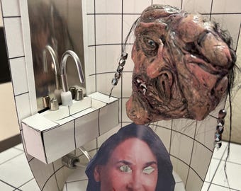May include: A diorama of a bathroom with a sink and mirror. A grotesque, fleshy creature with hair hangs above a woman's face with red lipstick and a wide smile. The creature has a beaded necklace.
