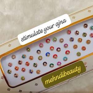 May include: A box of colorful, decorative, self-adhesive bindi stickers. The box has the text "stimulate your ajna" and "mehnidibeauty" printed on it.