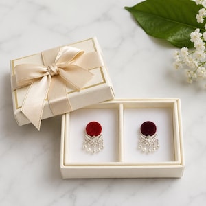 May include: A pair of circular stud earrings featuring red velvet tops and silver-toned metal bases with dangling white pearl accents, presented in a cream-colored gift box with a matching satin ribbon bow on the lid.