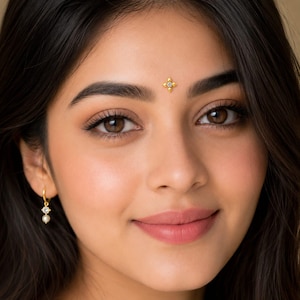 May include: A gold bindi featuring a four-pointed star design with a central clear stone, worn on the forehead, paired with a gold hoop earring that has a small clear stone and a tiny white pearl drop hanging from the bottom of the hoop.
