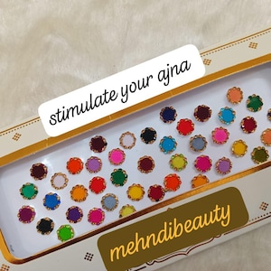 Può includere: Un foglio di adesivi rotondi colorati con bordatura dorata. Gli adesivi sono disposti a griglia e sono destinati ad essere applicati sulla pelle. Il foglio è etichettato "stimulate your ajna" e "mehnidibeauty".