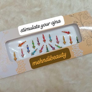 May include: A sheet of colorful, jewel-toned, decorative stickers in the shape of arrows. The stickers are packaged in a white box with the text "stimulate your ajna" and "mehnidibeauty".