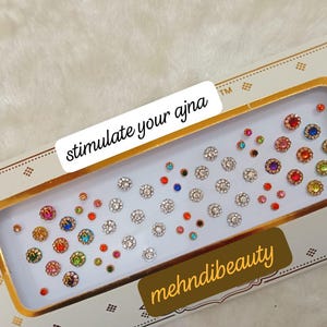 Puede incluir: Una hoja de gemas redondas, adhesivas y de colores para la cara o el cuerpo. Las gemas están dispuestas en filas sobre un fondo blanco con un borde dorado. El texto "stimulate your ajna" está impreso en el borde. El texto "mehnidibeauty" está impreso en la esquina inferior derecha.