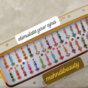 Può includere: Un foglio di gioielli per il corpo temporanei colorati, o bindis, in una scatola con bordo dorato. La scatola ha il testo "stimulate your ajna" e "mehnidibeauty" stampato su di essa.