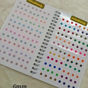 May include: A spiral-bound book with sheets of colorful, 6mm dot stickers. The stickers are arranged in rows and columns, with a variety of colors including pink, blue, green, and orange. The text "mehndi beauty" is printed on the cover.