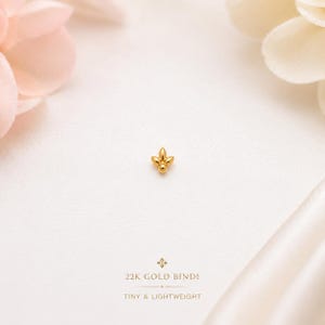 May include: A small, polished 22K gold bindi featuring a symmetrical, three-petaled floral design with a central rounded point. The text below the bindi reads 22K GOLD BINDI and TINY & LIGHTWEIGHT, accompanied by a small decorative star icon.
