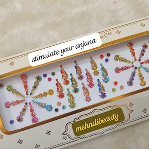 Può includere: Una scatola decorativa con una finestra bordata d'oro che mostra gioielli colorati per la fronte bindi. I gioielli sono disposti a forma di stella e presentano vari colori e forme. Il testo "stimulate your anjana" è visibile.