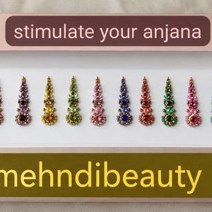 May include: A rectangular product box labelled "stimulate your anjana" and "mehndibeauty" containing twelve decorative bindi stickers. Each bindi features a teardrop shape with colourful embedded crystals in pink, blue, orange, purple, yellow, and green.