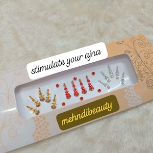 Può includere: Un pacchetto bianco e oro con il testo "stimulate your ajna" e "mehnidibeauty". Il pacchetto contiene un foglio di adesivi decorativi rossi e dorati.