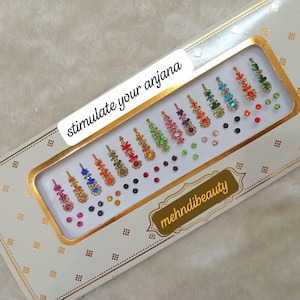 Puede incluir: Una caja decorativa que contiene una variedad de pegatinas de frente bindi de colores. Las pegatinas están dispuestas en filas y presentan varios colores y diseños de tonos joya. La caja tiene el texto "stimulate your anjana" y "mehdibeauty".