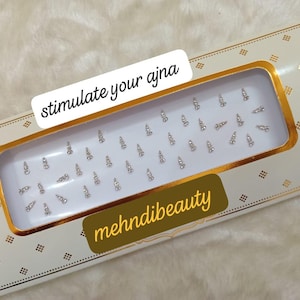Puede incluir: Una caja blanca y dorada con una ventana de plástico transparente que muestra una hoja de pequeños diamantes de imitación decorativos plateados. La caja tiene el texto "stimulate your ajna" y "mehnidibeauty" impreso en ella.