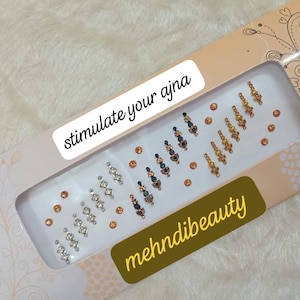 Può includere: Una confezione di adesivi bindi decorativi in una varietà di disegni e colori, tra cui argento, oro e blu scuro. La confezione presenta i testi "stimulate your ajna" e "mehdibeauty".