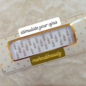 Puede incluir: Un paquete blanco y dorado de diamantes de imitación plateados, decorativos y autoadhesivos. El paquete tiene el texto "stimulate your ajna" y "mehnidibeauty" impreso en él.