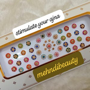 May include: A white box with a gold border containing a variety of colorful round stickers. The box has the text "stimulate your ajna" and "mehndibeauty" printed on it.