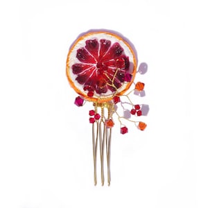 May include: A unique hair comb featuring a dried blood orange slice with a vibrant red center and orange rind. Adorned with red and orange crystals and delicate gold wire, this accessory adds a touch of nature-inspired elegance.