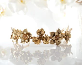 May include: A gold-colored floral crown with detailed flower and leaf designs. The crown is made of metal and reflects light, creating a shimmering effect. The flowers have a central gem. The crown is likely for a wedding or special occasion.