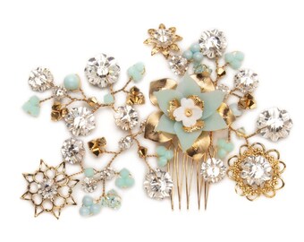 May include: A gold-toned hair comb with a floral design. The comb features a large, light blue flower with a white center, surrounded by clear crystals, gold leaves, and small, light blue beads. A starburst and a circular design with crystals are also included.