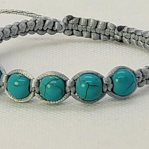 Op de afbeelding: Een grijs gevlochten armband met vijf ronde turquoise kralen.