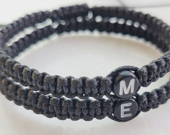 Puede incluir: Dos pulseras trenzadas negras con las letras "ME" en blanco sobre cuentas negras.