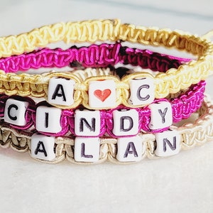May include: Three friendship bracelets with the names "Cindy" and "Alan" spelled out with white beads and a heart bead between the names "A" and "C". The bracelets are made with yellow, pink, and beige string.