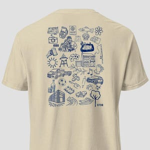 May include: Beige t-shirt with a navy blue doodle design on the back. The design includes illustrations of a camera, passport, fish, couple, house, car, and other objects. The year "1999" is also included in the design.