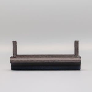 Puede incluir: Un objeto peque&ntilde;o, rectangular, gris y negro con dos postes verticales. El objeto parece estar hecho de materiales en capas. El fondo es de color gris claro liso.