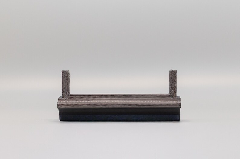 Puede incluir: Un objeto peque&ntilde;o, rectangular, gris y negro con dos postes verticales. El objeto parece estar hecho de materiales en capas. El fondo es de color gris claro liso.