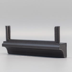 Puede incluir: Un objeto gris oscuro impreso en 3D con una base rectangular y dos postes verticales. El objeto tiene un dise&ntilde;o decorativo y curvo en la base. Los postes son texturizados. Probablemente sea un componente o una pieza.