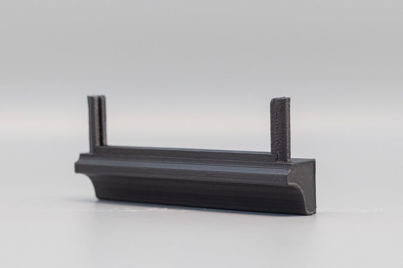 Puede incluir: Un objeto gris oscuro impreso en 3D con una base rectangular y dos postes verticales. El objeto tiene un dise&ntilde;o decorativo y curvo en la base. Los postes son texturizados. Probablemente sea un componente o una pieza.