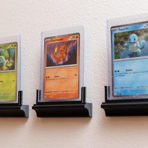 Puede incluir: Tres cartas coleccionables de Pok&eacute;mon enmarcadas, con Bulbasaur, Charmander y Squirtle, expuestas en estantes negros. Las cartas est&aacute;n en fundas protectoras, mostrando las ilustraciones coloridas y los detalles.