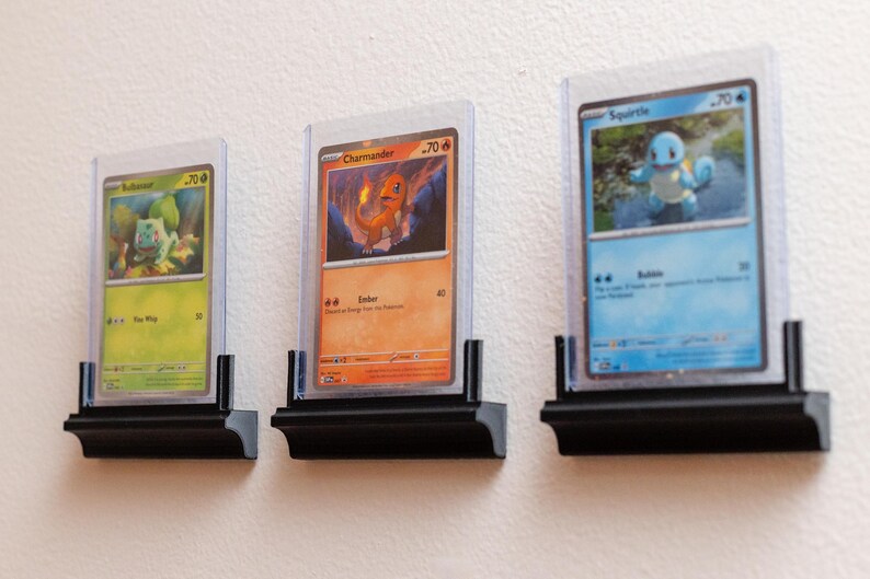 Puede incluir: Tres cartas coleccionables de Pok&eacute;mon enmarcadas, con Bulbasaur, Charmander y Squirtle, expuestas en estantes negros. Las cartas est&aacute;n en fundas protectoras, mostrando las ilustraciones coloridas y los detalles.