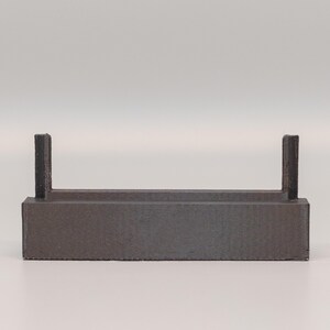 Puede incluir: Un objeto rectangular gris oscuro con dos soportes verticales. El objeto tiene una superficie texturizada y un dise&ntilde;o simple y minimalista. Podr&iacute;a ser un estante peque&ntilde;o o un expositor.