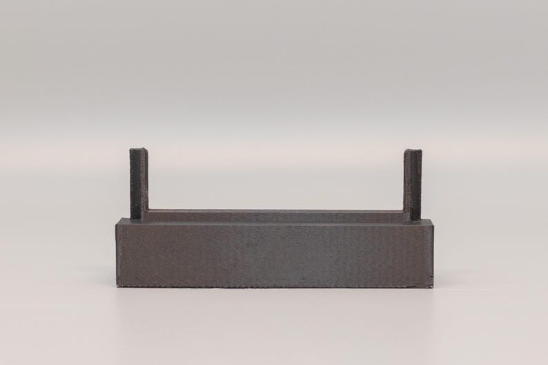 Puede incluir: Un objeto rectangular gris oscuro con dos soportes verticales. El objeto tiene una superficie texturizada y un dise&ntilde;o simple y minimalista. Podr&iacute;a ser un estante peque&ntilde;o o un expositor.