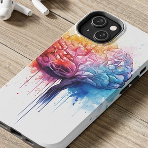 Puede incluir: Una funda de teléfono blanca con una pintura de acuarela colorida de un cerebro. El cerebro está pintado en tonos de rosa, azul, verde, amarillo y naranja. La funda del teléfono está sobre una superficie de madera.