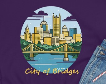 May include: Purple sweatshirt with a graphic of the Pittsburgh skyline and the text "City of Bridges". The sweatshirt is laid out on a white surface with a pair of black sneakers, a pair of blue jeans, and a beige beanie.